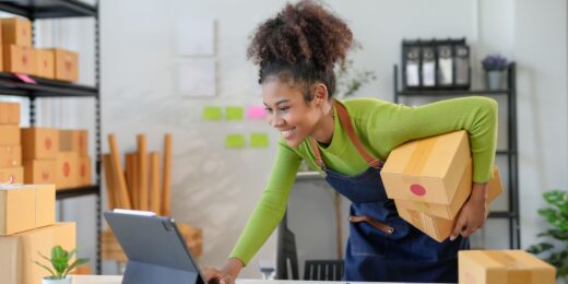 Motivated young entrepreneur managing her small business, holding packages and working on a tablet in a home office filled with shipping boxes