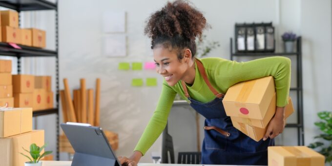 Motivated young entrepreneur managing her small business, holding packages and working on a tablet in a home office filled with shipping boxes