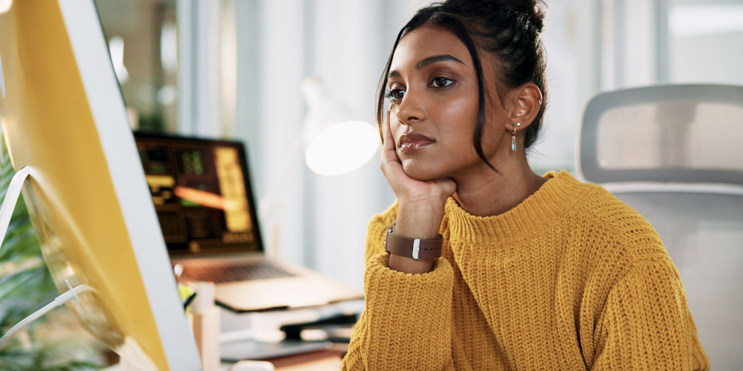 Woman in a yellow sweater sitting at a desk, looking thoughtfully at a computer screen, procrastinating, with a laptop and office supplies in the background
