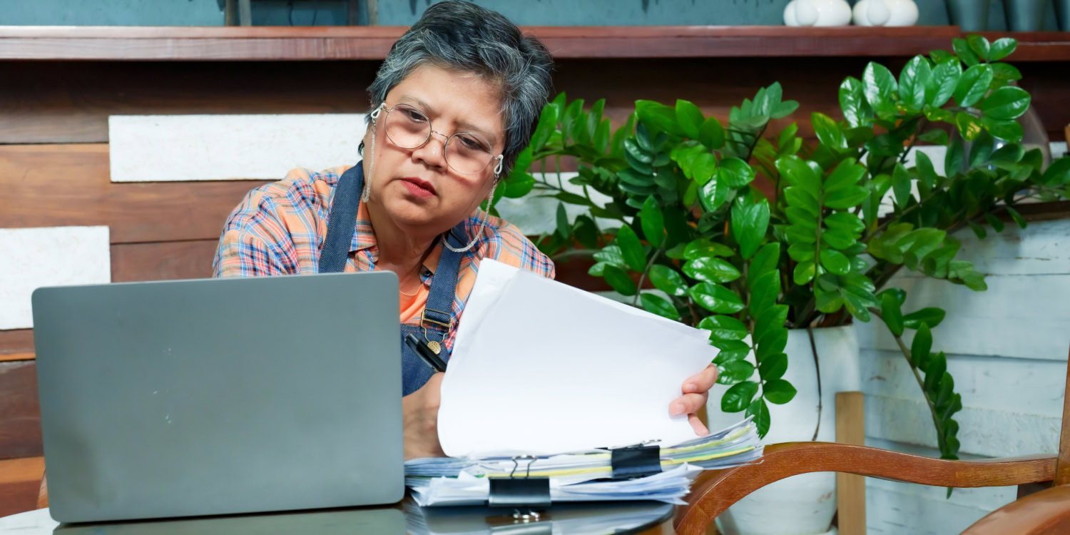 Small business owner facing a challenge, reviewing documents and working on a laptop at a cluttered desk.