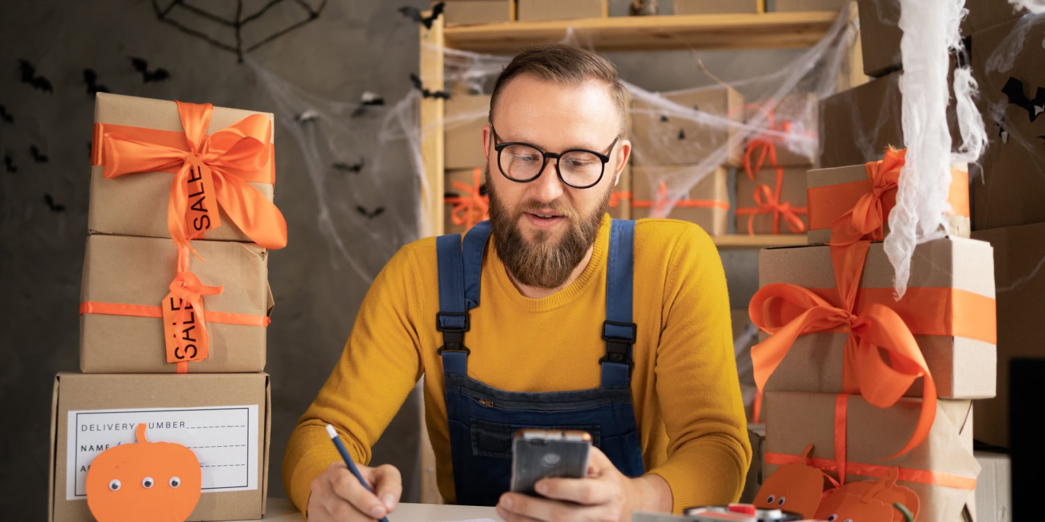 Small business owner surrounded by Halloween-themed packages with orange ribbons, working on a Halloween marketing idea while using a smartphone and writing notes. Decorations include cobwebs, bats, and pumpkins