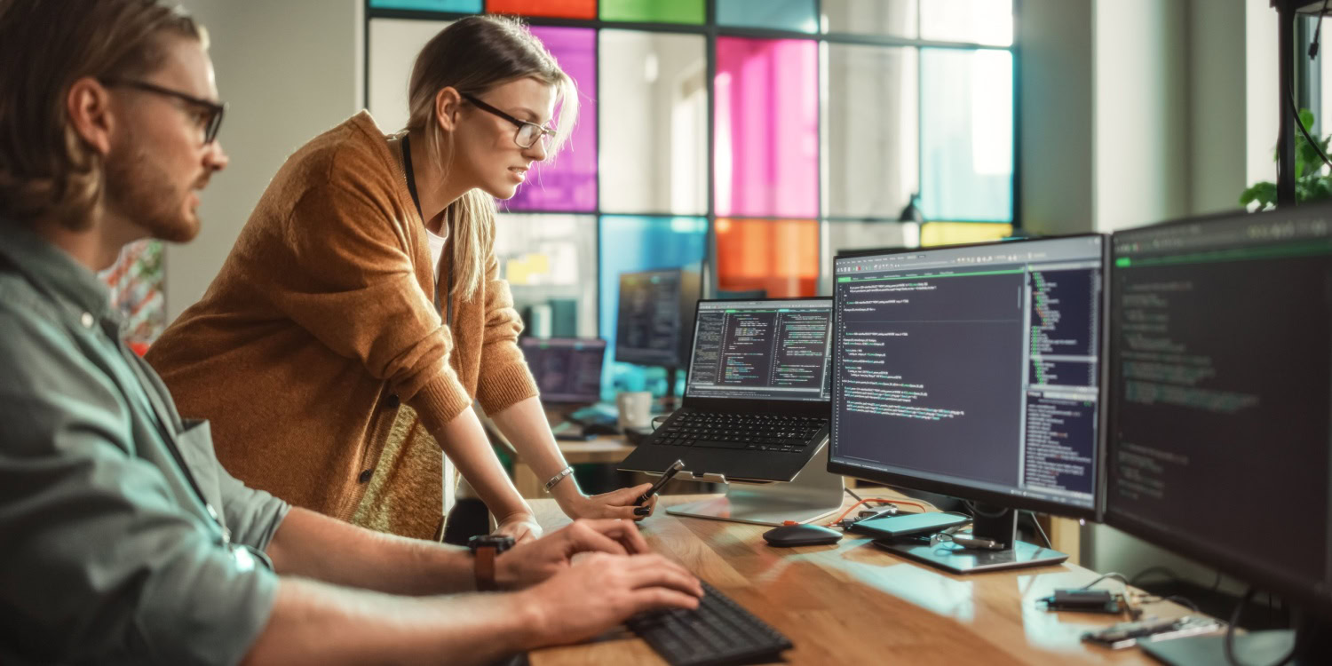Two professionals working on multiple computer screens with code displayed, representing teamwork and digital transformation in a modern office setting