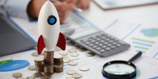 Toy rocket standing on stacks of coins surrounded by charts, calculator, and magnifying glass, symbolising pre-seed funding for a startup