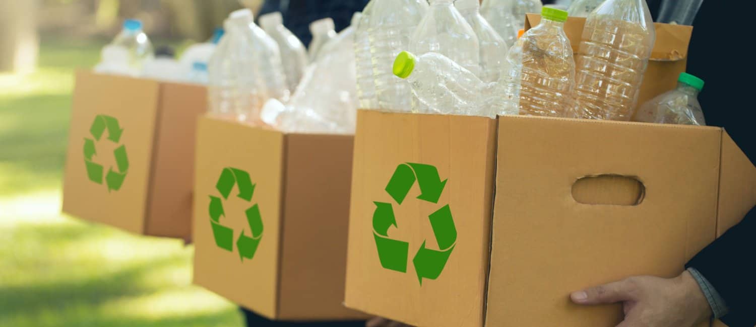 Three people, each holding a cardboard box with a recycling symbol on the front. Boxes are filled with plastic bottles, ready to be recycled.