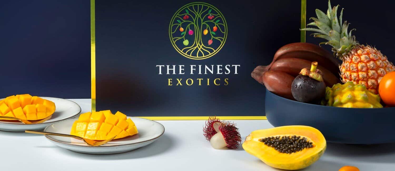 Display of The Finest Exotics packaging with exotic fruits artfully displayed in the foreground on plates and in a fruit bowl