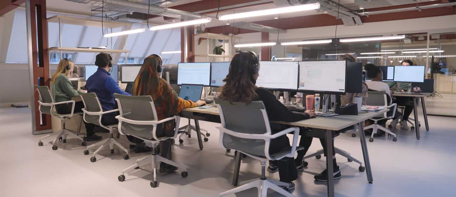 A modern office space with several people working at desks, focused on multiple computer screens, wearing headphones.