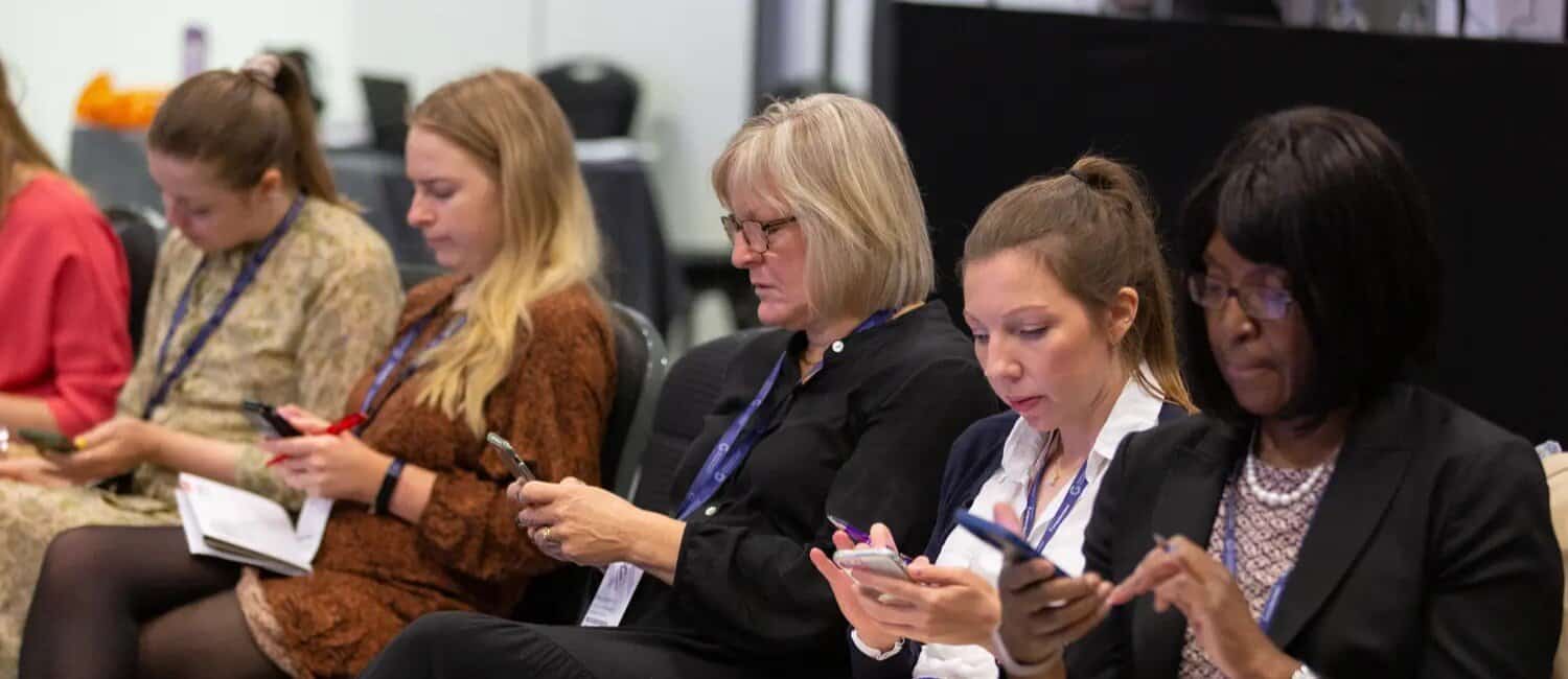 A group of women sits in a conference setting, engaged with their smartphones and notebooks.