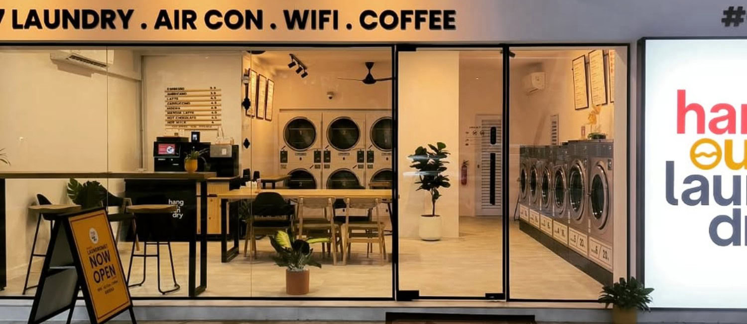 Hangout Laundry in Bedok, Singapore: a modern laundromat with washing machines, dryers, coffee counter, and communal workspace seating behind a glass storefront.
