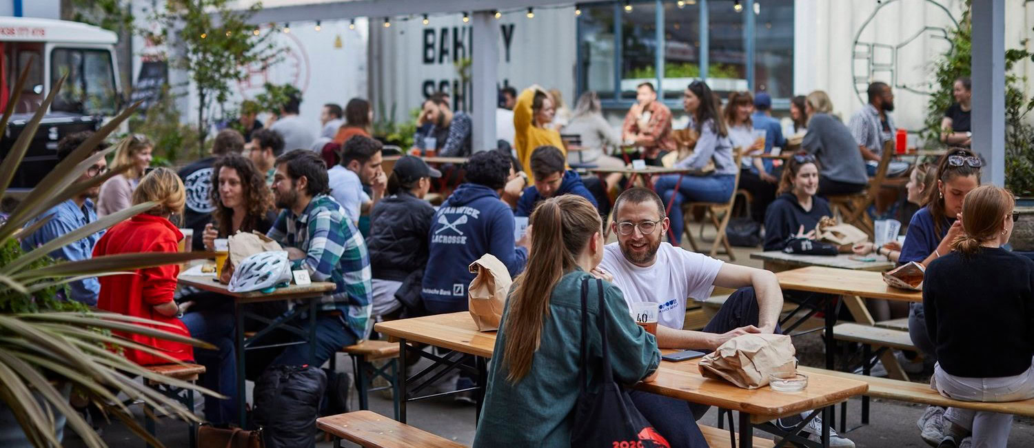 Outdoor seating at The Dusty Knuckle bakery and café in Dalston, London, with people chatting and eating at wooden benches in a lively courtyard.