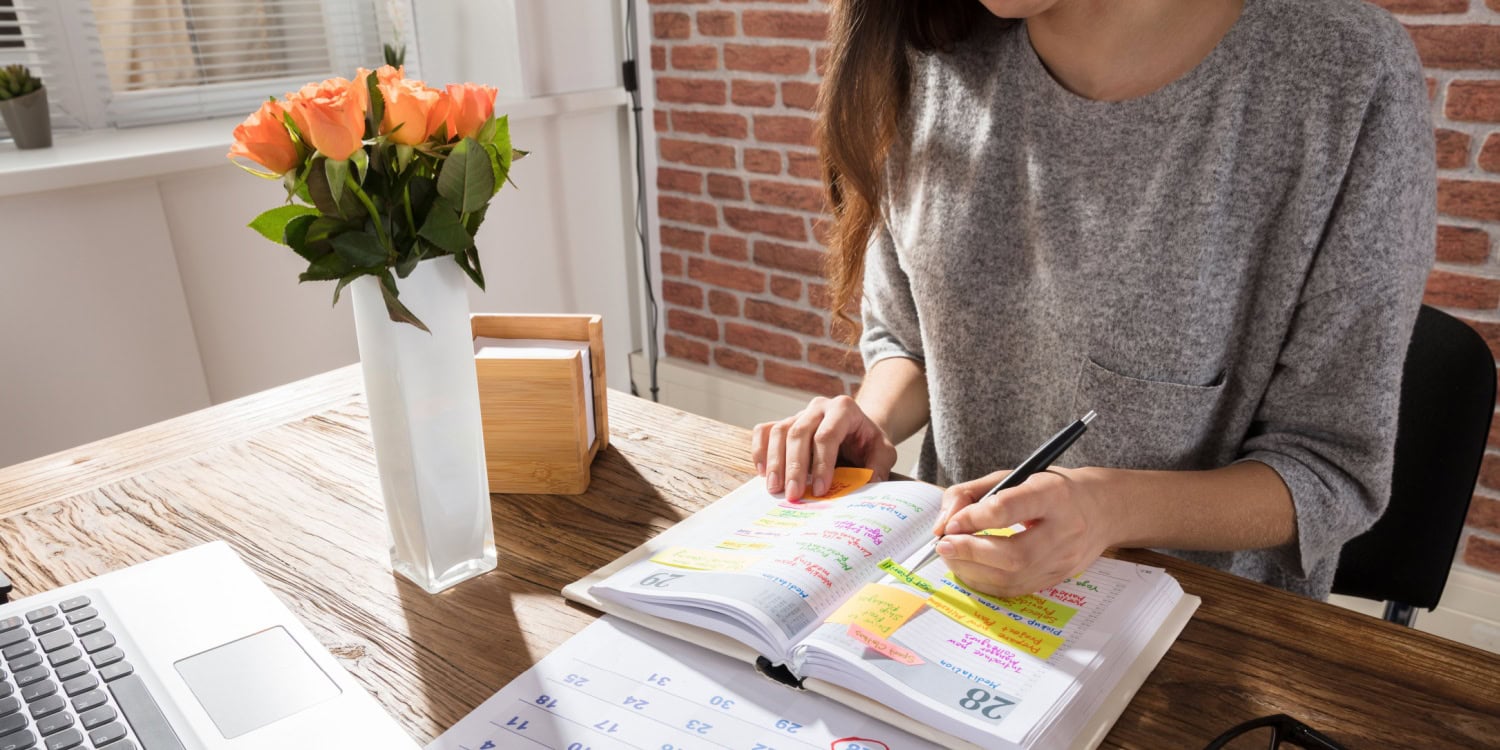 Woman highlighting dates in a planner at a desk with a laptop and flowers, considering the best time of year to start a business