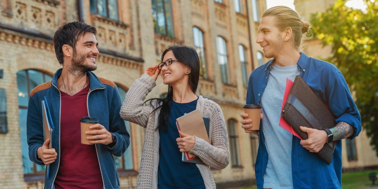 Three young international students walking and chatting outside a university-style brick building, holding notebooks and takeaway coffee cups.