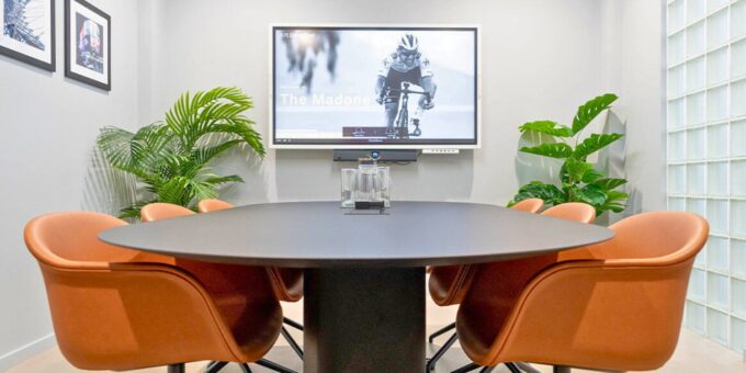 Modern meeting room with a round table, orange chairs, a screen displaying a cyclist, and plants in the background.