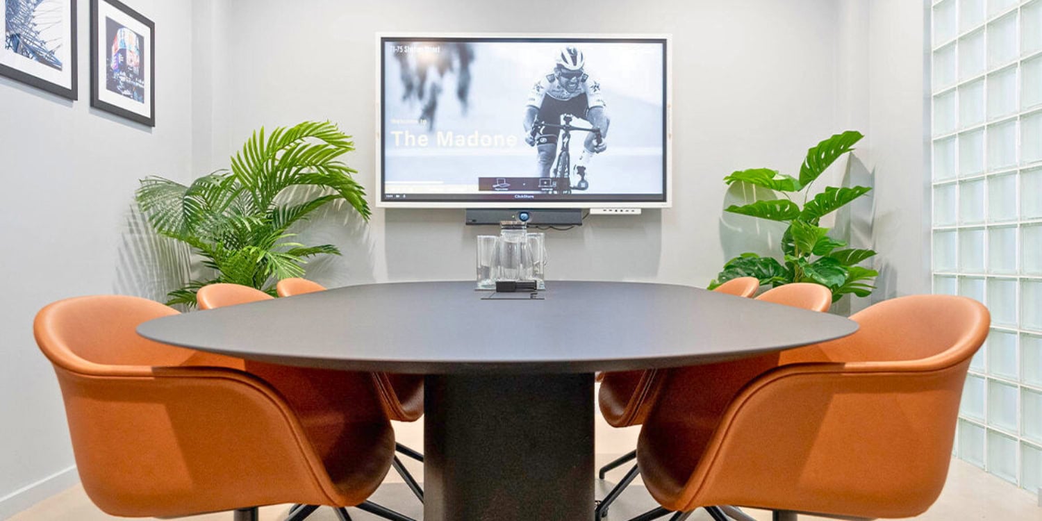 Modern meeting room with a round table, orange chairs, a screen displaying a cyclist, and plants in the background.