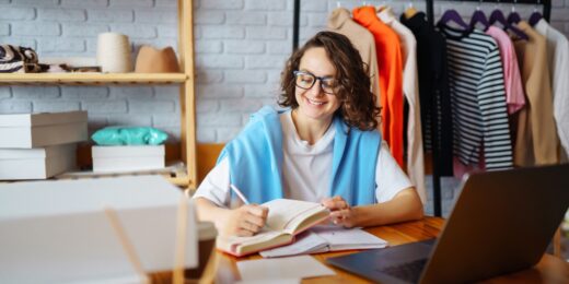 Woman smiling while writing in a notebook, seated at a laptop in a clothing business workspace.