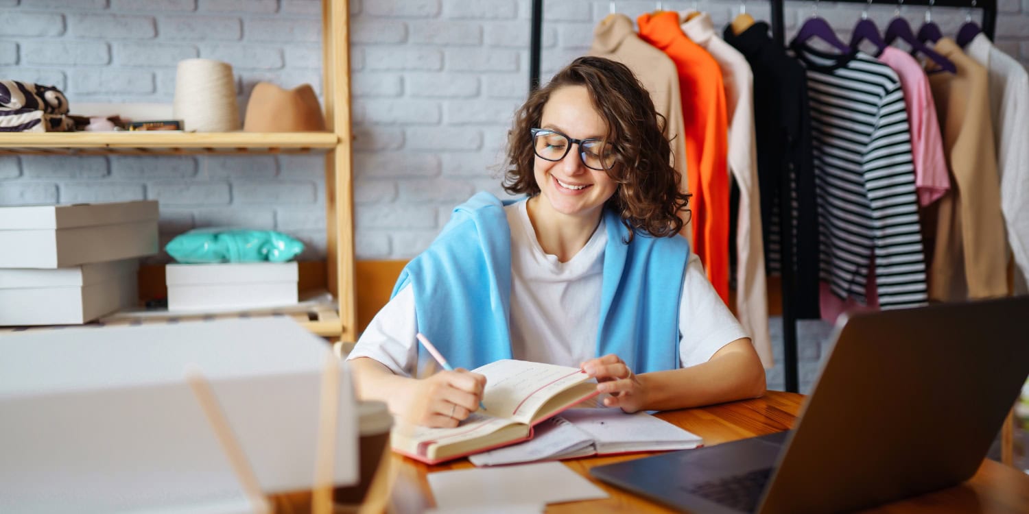 Woman smiling while writing in a notebook, seated at a laptop in a clothing business workspace.