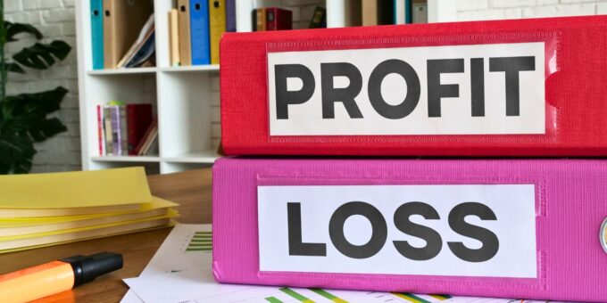 Two binders labeled "PROFIT" and "LOSS" stacked on a wooden desk, with yellow folders and financial documents in the background, in a bright office environment.