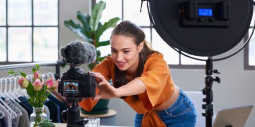 Young female content creator adjusting a DSLR camera on a tripod with a ring light in a bright home studio, with a laptop on the desk.