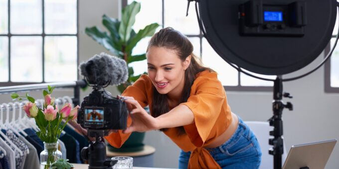 Young female content creator adjusting a DSLR camera on a tripod with a ring light in a bright home studio, with a laptop on the desk.