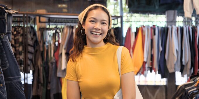 Young woman in a yellow top and headband smiles while standing in a clothing store with racks of garments visible behind her.