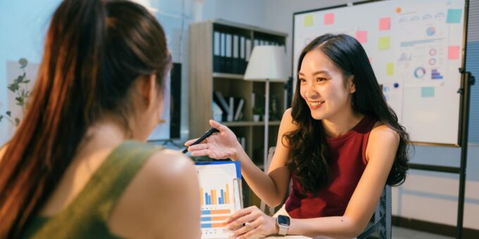 Two colleagues in an office meeting, one woman smiling and pointing to charts on a clipboard with a whiteboard in the background.