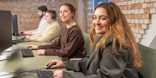 Four colleagues seated at computers in an open-plan office, two women in the foreground smiling at the camera while two coworkers work in the background.