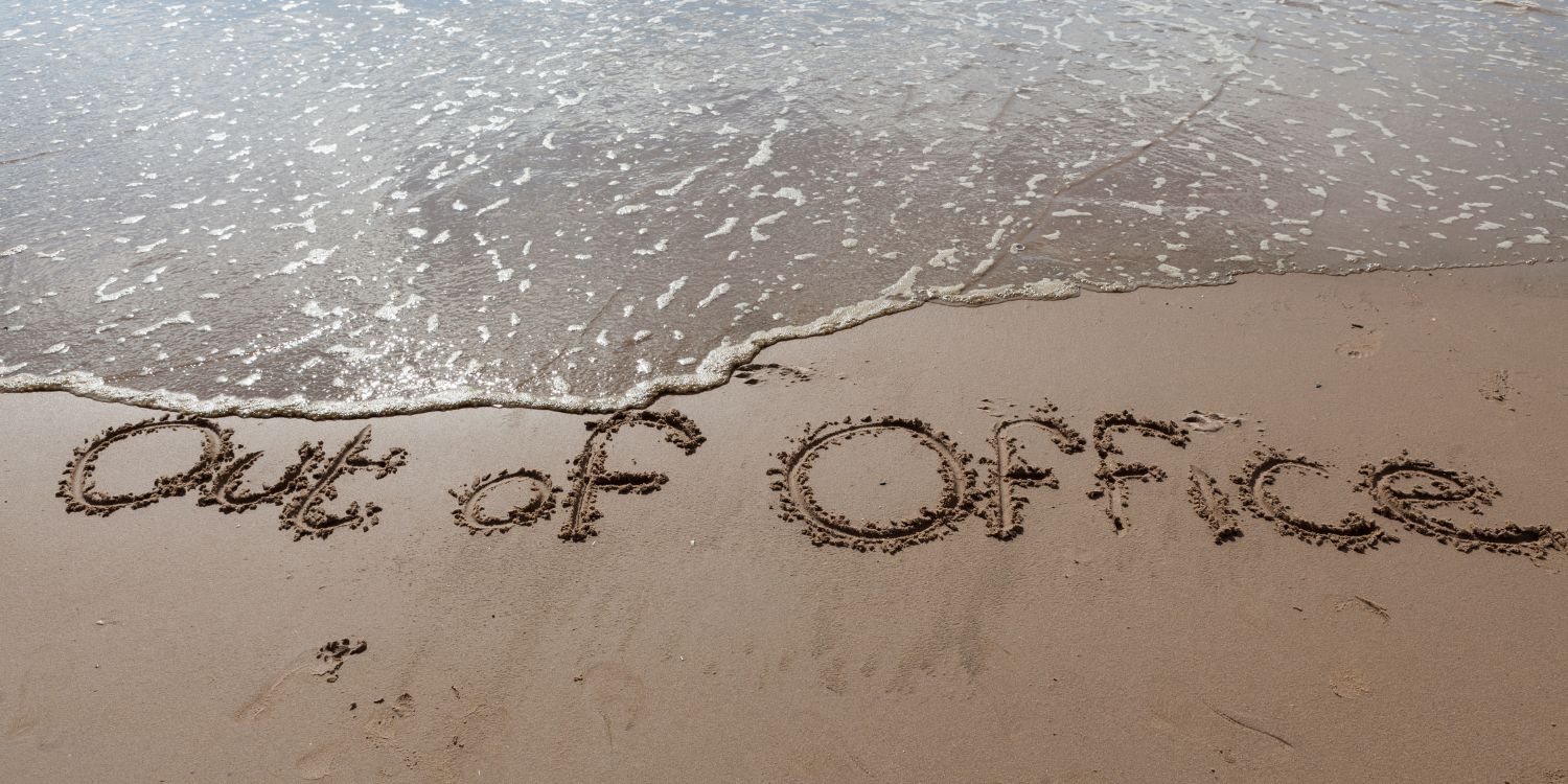 The words 'out of office' drawn in the sand on a beach, next to the ocean.