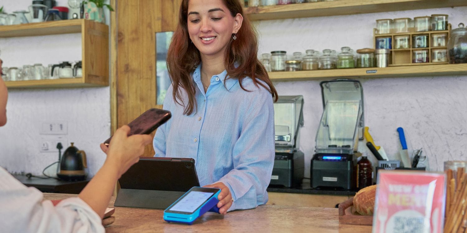 A small business owner standing behind the counter of a cafe, holding a card reader, while a customer taps her phone to pay.
