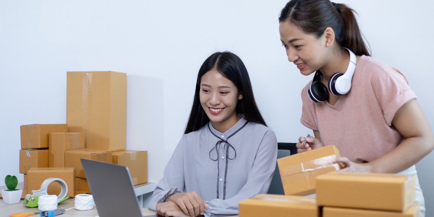 Two small business owners packing customer orders at a desk surrounded by cardboard boxes, reflecting the early stages of finding and fulfilling a business's first customers.