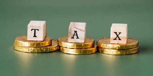 Wooden letter blocks spelling "TAX", each resting on a stack of gold coins against a green background, illustrating the new tax year changes.