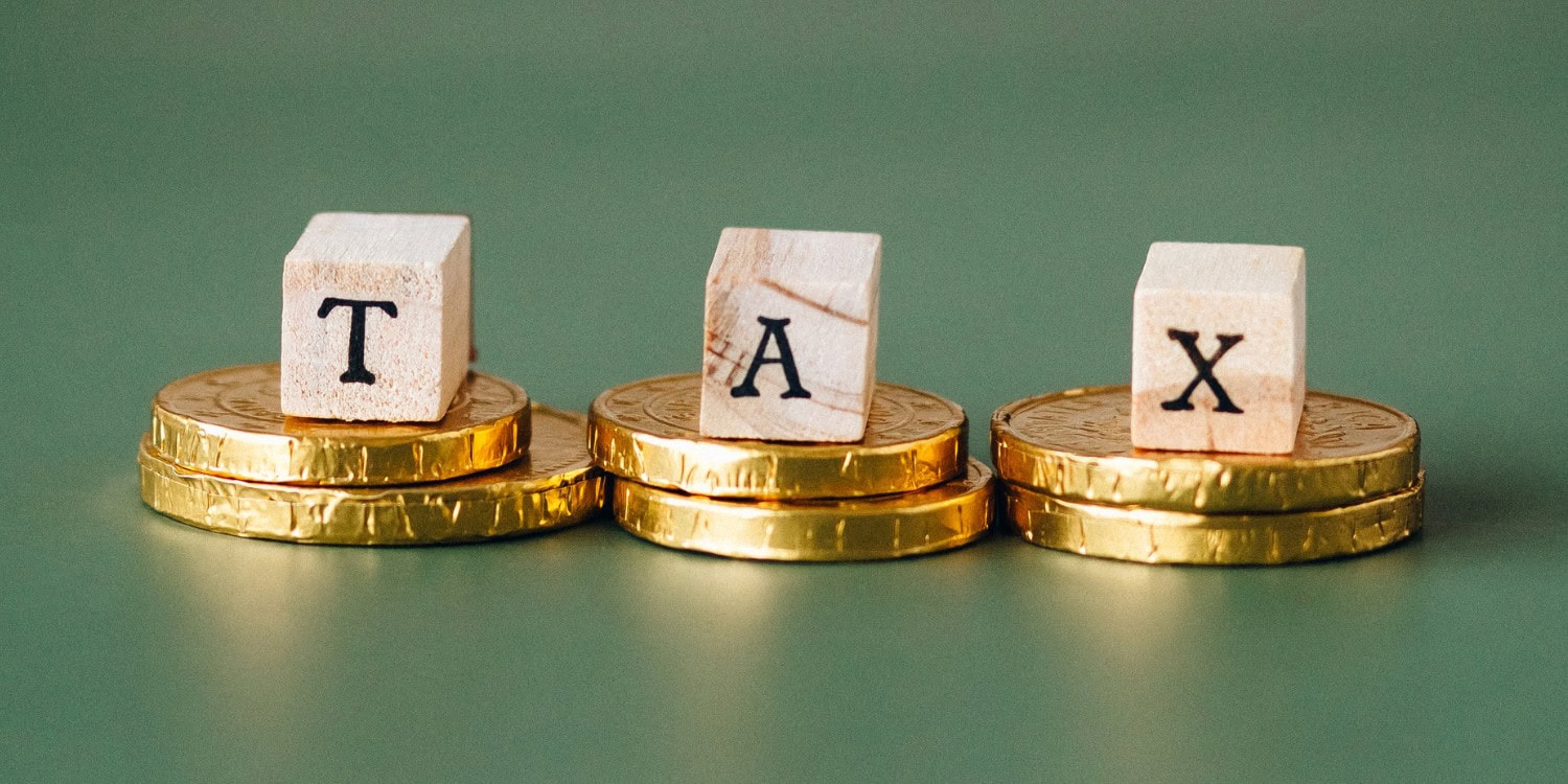 Wooden letter blocks spelling "TAX", each resting on a stack of gold coins against a green background, illustrating the new tax year changes.