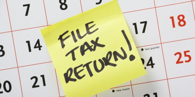 A yellow sticky note reading "File tax return!" stuck on a calendar page, as a reminder of upcoming tax dates and deadlines.